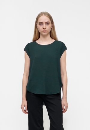 Jeune femme aux cheveux blonds raides portant un chemisier vert foncé à manches courtes et un pantalon noir sur un fond blanc uni.