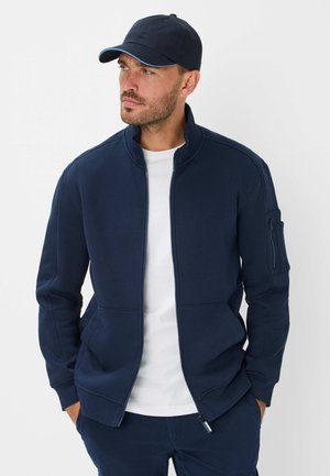 Marineblaue Zip-Jacke mit hohem Kragen, seitlichen Taschen und einer Armtasche. Getragen über einem weißen T-Shirt, kombiniert mit einer passenden Mütze.