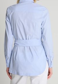 Camisa de botones azul claro con cintura ceñida, cuenta con cuello tipo solapa, mangas largas y un dobladillo curvado. Tejido suave y ligero.