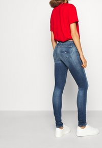 Jeans ajustados de mezclilla azul oscuro con bolsillos traseros cosidos. Combinados con una camisa roja de manga corta y zapatillas blancas. Textura suave.