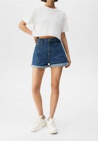 Pantalones cortos de mezclilla con dobladillos enrollados y cintura alta, combinados con una camiseta corta blanca y holgada. El modelo lleva zapatillas blancas.