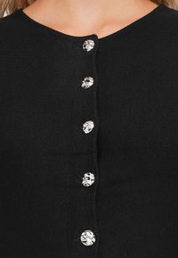 Cardigan noir avec un col rond, texture côtelée et six boutons décoratifs argentés avec une surface à motifs.