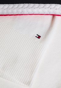 Zbliżenie na biały dziany ubiór z detalem prążkowanym, oplecionym obszyciem, czerwoną linią akcentową oraz małym prostokątnym logo w kolorach czarnym, białym i czerwonym.