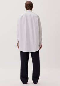 Camicia bianca a maniche lunghe con righe blu verticali, caratterizzata da una vestibilità rilassata, fessure laterali e un orlo arrotondato. Abbinata a pantaloni scuri.