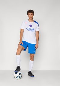T-shirt de sport blanc avec des accents bleus, logo "QATAR AIRWAYS", associé à un short bleu, des chaussettes blanches, des crampons noirs et un ballon de soccer.