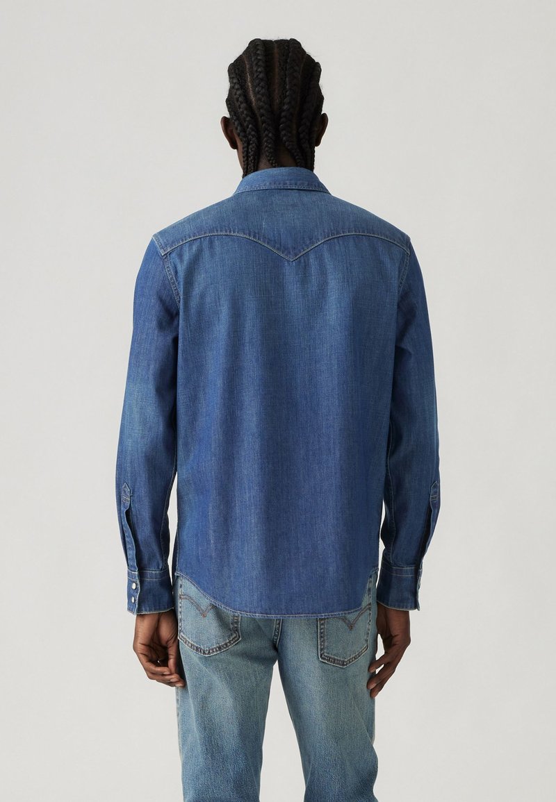 Camisa de mezclilla azul con un yugo puntiagudo, mangas largas y puños con botones a presión. Presenta un dobladillo ligeramente curvado y una textura suave.