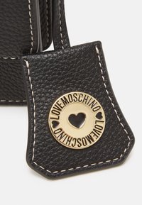 Svart texturerad läderetikett med krämfärgad sömnad, med en rund guldlogotyp präglad med "LOVE MOSCHINO" och ett hjärta uthugget.