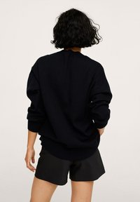 Marineblauer Strickpullover mit lockerer Passform, überschnittenen Schultern und gerippten Bündchen. Kombiniert mit schwarzen Shorts, was einen lässigen, geschichteten Look zeigt.