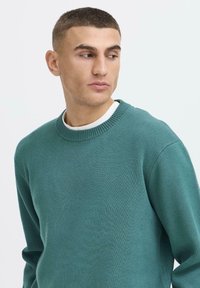 Jonge man met kort donker haar, gekleed in een teal ronde hals trui over een wit shirt, kijkt naar links tegen een effen witte achtergrond.