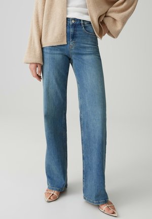 Frau trägt hellblaue Jeans mit hoher Taille und geradem Bein, beigen offenen Cardigan, weißes Top und beigefarbene Riemchen-Sandaletten vor einfarbigem Hintergrund.