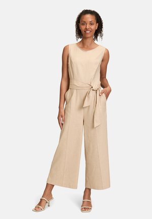 Salopette in lino beige con busto aderente, pantaloni a gamba larga e cintura legata. Il modello si trova in posizione con una mano in tasca.