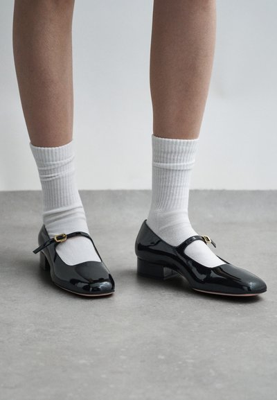 Des chaussures Mary Jane en cuir verni noir avec un bout carré, un petit talon et une bride à boucle dorée, portées avec des chaussettes blanches à côtes.