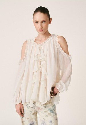 Frau mit zurückgestrichenen Haaren, die eine weiße, mit Rüschen verzierte, schulterfreie Bluse und hell gemusterte Blumenhose trägt, posiert mit einer Hand in der Hüfte.