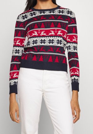 Vrouw draagt een feestelijke gebreide trui met rode, marineblauwe en witte kerstpatronen van rendieren, bomen en sneeuwvlokken, gecombineerd met een witte broek.