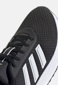Gros plan d'une chaussure de sport noire avec un logo blanc à trois bandes, des lacets noirs et une tige en mesh respirant sur une semelle blanche.