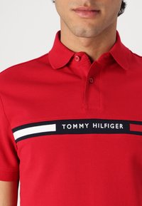 Rød polo skjorte lavet af struktureret bomuld, med et sort bånd med "TOMMY HILFIGER" logoet og hvide og sorte stribede detaljer.