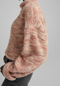 Różowy i kremowy puszysty sweter z wysokim kołnierzem, z teksturowanym wzorem i oversize'owymi rękawami, w parze z ciemnymi dżinsami.