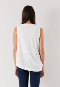 Blusa bianca senza maniche con orlo curvo e cucitura orizzontale. Realizzata in tessuto morbido, presenta un piccolo etichetta rosa sul retro.