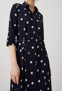 Donna che indossa un abito blu navy con pois bianchi, con bottoni sulla parte davanti, maniche a tre quarti e una cintura abbinata in vita.