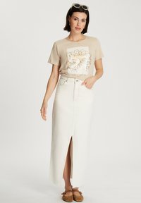 Beżowa koszulka graficzna z kwiatowym wzorem, w połączeniu z długą białą spódnicą dżinsową z rozcięciem z przodu. Model nosi brązowe pantofle na slip-on.