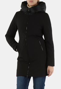 Cappotto nero fino al ginocchio con cappuccio imbottito e lucido, chiusura frontale con zip, tasche laterali e trama liscia. Indossato con jeans blu.