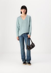 Pull en tricot texturé bleu clair avec un col en V ; porté avec un jean évasé bleu. Un sac à main foncé est tenu par le modèle. Des baskets noires complètent le look.