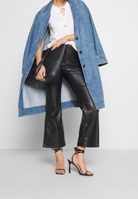 Denim coat with oversized fit, white top with tie neck, black leather flared pants, and strappy black high heels. Black handbag held in hand.