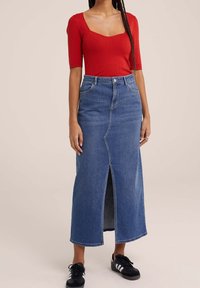 Rotes, geripptes Kurzarmoberteil kombiniert mit einem hoch taillierten blauen Jeans-Maxikleid mit einem vorderen Schlitz und getragen mit schwarzen Sportschuhen.