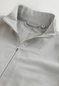 Gai pelēks laika jaku ar rāvējslēdzēju, augstu apkakli, gludu tekstūru un metāla rāvējslēdzēju. Auduma birka norāda izmēru M.