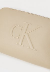 Beige läderplånbok med präglad "CK"-logotyp, slät yta, rundade kanter och kontrasterande sömmar längs omkretsen.