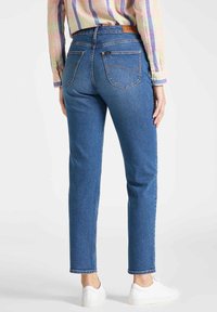 Jeans de mezclilla en azul oscuro con corte recto, diseño de cinco bolsillos y costuras visibles. Llevados con zapatillas blancas y una camisa a cuadros.