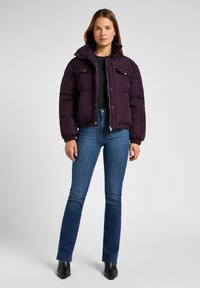 Chaqueta de plumas púrpura con botones a presión, dos bolsillos en el pecho y puños elásticos. Combinada con jeans azules y botas negras de puntera afilada.