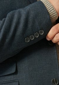 Manica di un cappotto blu navy con un tessuto a trama e quattro bottoni neri. Sotto, un maglione a maglia color marrone chiaro è parzialmente visibile.