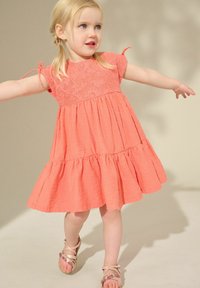 Korallenfarbenes Kleid mit Spitzenoberteil und kurzen Ärmeln, ausgestattet mit einem ausgestellten Rock mit rüschengesäumtem Saum. Kombiniert mit dekorativen Sandalen.