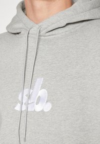 Acercamiento de una sudadera gris claro con un logo estilizado en blanco "slb." bordado en el pecho y cordones a juego.