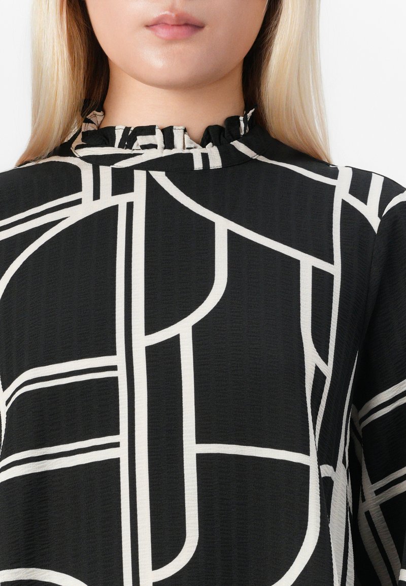 Mujer vistiendo una camiseta negra con líneas geométricas abstractas en blanco y un pequeño escote con volantes.