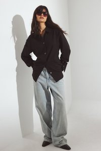 Svart skjorta med knappar och ljusblå vida jeans. Svarta spetsiga skor. Modell som bär stora mörka solglasögon, står mot en ljus bakgrund.