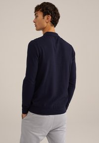 Polo de punto azul marino con puños y dobladillo acanalados, con un cuello clásico, visto desde la parte de atrás. Combinado con pantalones gris claro.
