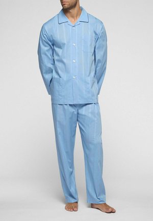 Uomo in piedi a piedi nudi che indossa una camicia da pigiama azzurro chiaro con maniche lunghe e bottoni e pantaloni da pigiama larghi abbinati su sfondo neutro.