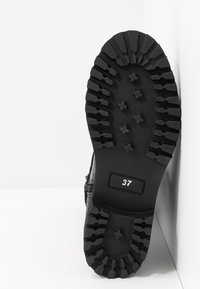 Semelle de botte noire avec des crampons profonds et le numéro de taille 37 visible, positionnée debout contre un coin blanc.