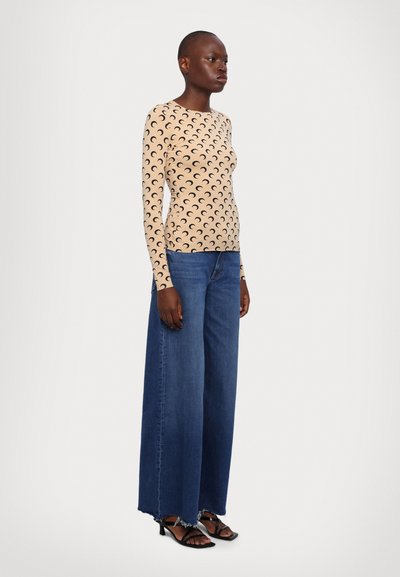 Blusa beige de manga larga con patrones de media luna, combinada con jeans azules holgados. Sandalias de tacón bajo completan el atuendo.