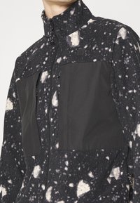 Uomo che indossa una giacca di pile a macchie bianche e nere con tasche frontali nere uniformi e zip, visibile dalla gola alla vita.