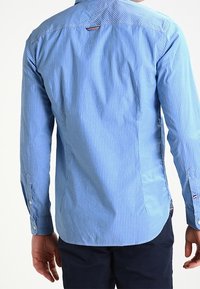 Modrá pruhovaná košile na knoflíky s límečkem, dlouhými rukávy a zakulaceným lemem. Má kontrastní šití a detail loga na zadní straně.