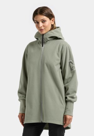 Frau trägt eine hellgrüne Fleecejacke mit Reißverschluss, hohem Kragen und Kapuze, steht vor einem schlichten hellen Hintergrund.