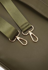 Olivgrüner Schulterriemen mit strukturiertem Finish, ausgestattet mit goldenen Metallclips zur Befestigung, vor einem glatten, dunkelgrünen Hintergrund.