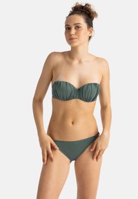 Žalias bandeau tipo maudymosi kostiumėlio viršus su susiraukšlėjusiu audiniu ir atitinkamu žemu rasu. Yra dekoratyvinis akcentas šone maudymosi kostiumėlio apačios.