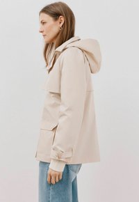 Beige vattentät jacka med huva, framflik och sidofickor. Har en figursydd design med uppvikt ärm över en lätt tröja.