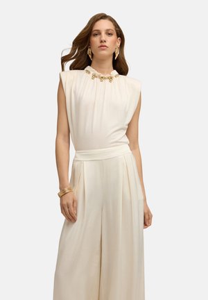 Vrouw draagt een mouwloos wit topje met gouden kraaldetail aan de hals, wijde witte broek, gouden oorbellen en armbanden, staand tegen een witte achtergrond.