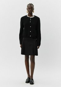 Črna pletena jakna z belim ovratnikom, z velikimi gumbi in dvema žepoma, kombinirana s črno nabranih krilom in loafersi.