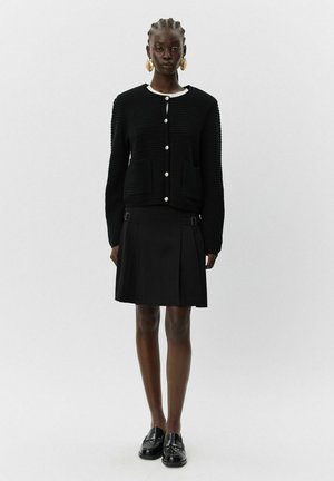 Svart stickad kofta med vit krage, prydd med stora knappar och två fickor, tillsammans med en svart plisserad kjol och loafers.